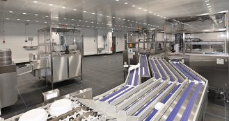 Automatisierung bei Vivantes: Der Spülbereich im Vivantes Speisenverteilzentrum. Das gespülte Geschirr läuft sortenrein aus, hin zu den Greifarmen, die alles hinter einer Schutzwand abnehmen und in Stapler einbringen.